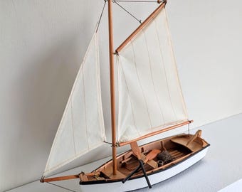 May include: A detailed model sailboat with a white hull and sails. The wooden mast and boom support the sails, and oars are visible inside the boat. The boat is a miniature replica of a classic sailing vessel.
