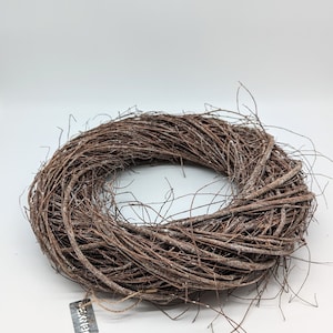 Könnte beinhalten: Ein kreisförmiger Kranz aus miteinander verflochtenen braunen Zweigen. Der Kranz hat ein natürliches, rustikales Aussehen, mit einem Etikett "Exkier", das mit Bindfaden befestigt ist. Die Zweige sind dicht gepackt und erzeugen ein volles, strukturiertes Aussehen, ideal für die saisonale Dekoration.