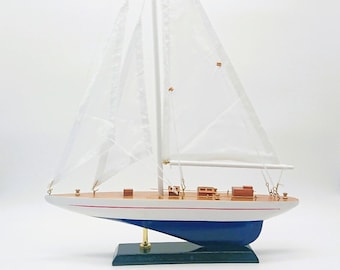 Puede incluir: Un modelo de velero de madera blanco con detalles azules y rojos. Las velas son blancas y el barco está sobre una base verde.