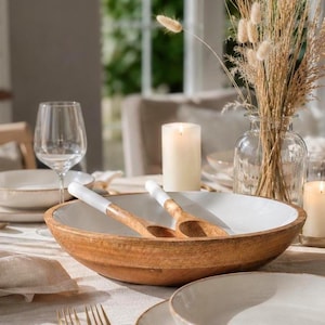 Puede incluir: Un cuenco de madera con interior blanco y dos cucharas de servir de madera con mangos blancos. El cuenco está rodeado de platos, velas y un jarrón de cristal con flores secas, creando un ambiente de mesa cálido y acogedor.