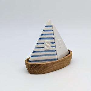 Puede incluir: Un juego de salero y pimentero de temática náutica. Los saleros en forma de vela son blancos con rayas azules y las letras "S" y "P". La base tiene forma de barco de madera.