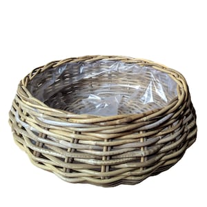 Puede incluir: Una cesta redonda de mimbre con un forro de plástico transparente. La cesta está hecha de mimbre de color marrón claro y gris, con un patrón tejido apretado. La cesta está diseñada para guardar plantas u otros artículos.