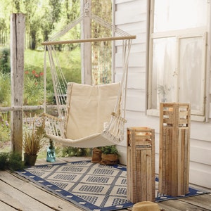 Könnte beinhalten: Ein weißer Hängesessel mit einem beigefarbenen Kissen, zwei Kerzenständern aus Holz und einem blau-weißen Teppich auf einer Holzveranda.