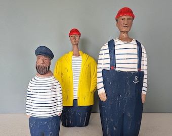 May include: Three wooden sailor figurines. The figures wear blue and white striped shirts, blue pants, and red hats. One figure has a yellow jacket. The tallest figure has blue overalls with an anchor detail. The figures are on a gray background.