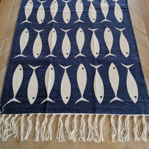 Könnte beinhalten: Ein rechteckiger marineblauer Teppich mit einem sich wiederholenden Muster aus cremefarbenen Fischen. Der Teppich hat einen Fransenbesatz am unteren Rand. Der Teppich besteht aus gewebtem Material und eignet sich für ein Wohn- oder Schlafzimmer.