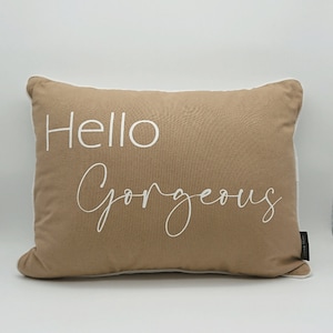 May include: A rectangular tan pillow with white trim and the words "Hello Gorgeous" in white script. The pillow has a soft, inviting appearance, suitable for home decor.