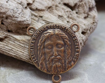 May include: A round bronze-toned metal connector featuring a relief of a bearded face in the centre. The text "ILLUMINA DOMINE VULTUM TUUM SUPER NOS" is embossed around the perimeter. It includes three small circular loops for attachment.