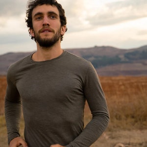Könnte beinhalten: Ein Mann in einem langärmeligen, dunkelgrünen T-Shirt läuft durch ein Feld. Das Feld ist braun und trocken, mit Hügeln im Hintergrund.