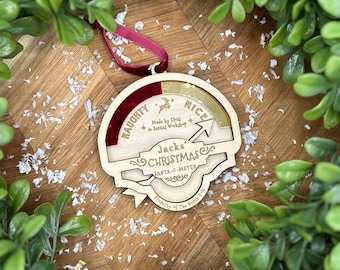 May include: A wooden Christmas ornament with a red, gold, and white color scheme. It features the words "Naughty or Nice" and "Jack's Christmas Santa-C-Meter" with a red ribbon.