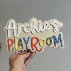 May include: A wooden sign that reads "Archie's PLAYROOM" in layered, colorful lettering. The word "Archie's" is in white, cursive font, while "PLAYROOM" is in various colors. The sign is held by a person.