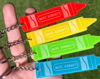 May include: Four colorful crayon-shaped keychains in red, yellow, green, and blue. Each keychain has the text "MISS ROBERTS" in a white oval. The keychains are attached to silver keyrings and chains.