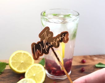 May include: A gold-colored acrylic cocktail stirrer with the text "The Wilkinsons" in a cursive font. The stirrer is in a glass of a refreshing drink with ice, lemon, and pomegranate seeds.
