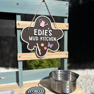 May include: A blue and wood children's play kitchen with a black sign that says "Edie's Mud Kitchen" with a pink watering can, a butterfly, and a flower. The kitchen has a stainless steel pot and three black and white stovetop burners.