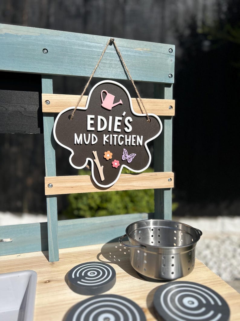 May include: A blue and wood children's play kitchen with a black sign that says "Edie's Mud Kitchen" with a pink watering can, a butterfly, and a flower. The kitchen has a stainless steel pot and three black and white stovetop burners.