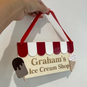 May include: A wooden sign with the text "Graham's Ice Cream Shop" and decorative ice cream cone and popsicle accents. The sign has a red and white awning design and a red ribbon for hanging.