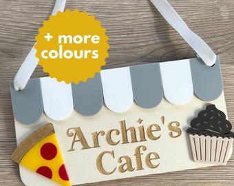 May include: Wooden cafe sign with a white ribbon for hanging. The sign features a gray and white awning design, the text "Archie's Cafe", a pizza slice, and a cupcake. A yellow circle with the text "+ more colours" is also present.