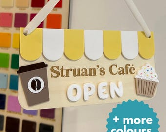 May include: A wooden cafe sign with a yellow and white awning design. The sign features the text "Struan's Cafe OPEN", a coffee cup, and a cupcake. A white ribbon is attached for hanging.