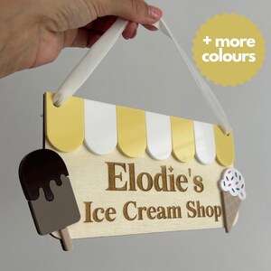 May include: Wooden sign for "Elodie's Ice Cream Shop" with a yellow and white awning design. Features an ice cream bar and cone illustration. Includes a white ribbon for hanging and a yellow circle with "+ more colours" text.