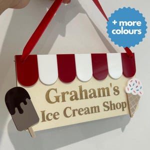 May include: Wooden sign for "Graham's Ice Cream Shop" with red and white awning design. Features an ice cream cone and popsicle. Includes a red ribbon for hanging. The sign is cream-colored with gold lettering.