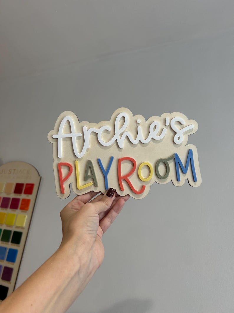 May include: A wooden sign that reads "Archie's PLAYROOM" in layered, colorful lettering. The word "Archie's" is in white, cursive font, while "PLAYROOM" is in various colors. The sign is held by a person.