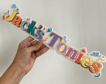 May include: A colorful sign that reads "Jack's Tonies" in a playful font. The letters are in various colors, including yellow, teal, purple, and orange, with gold trim. Confetti and decorative swirls surround the text.