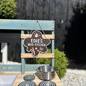 May include: A blue and brown wooden play kitchen with a chalkboard sign that says "Edie's Mud Kitchen". The kitchen has a sink, a stovetop with three burner knobs, and a pot.