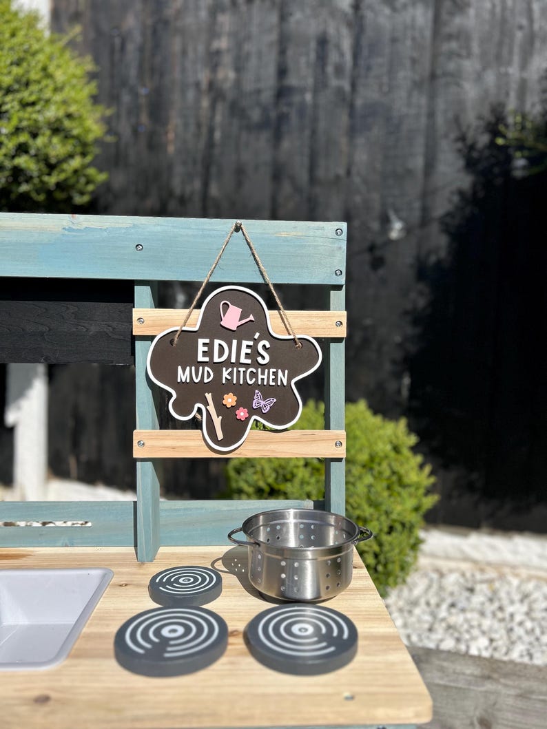 May include: A blue and brown wooden play kitchen with a chalkboard sign that says "Edie's Mud Kitchen". The kitchen has a sink, a stovetop with three burner knobs, and a pot.