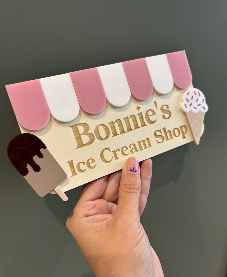 May include: A wooden sign for "Bonnie's Ice Cream Shop" with a pink and white awning design. The sign features a popsicle and an ice cream cone with sprinkles. The text is engraved in a classic font.