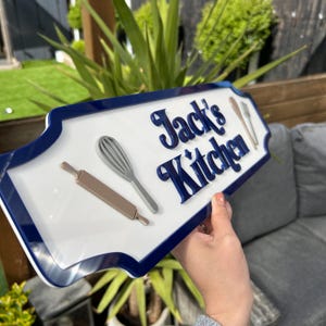 May include: White sign with a blue border and the text "Jack's Kitchen" in blue raised letters. The sign has a rolling pin and a whisk on it.