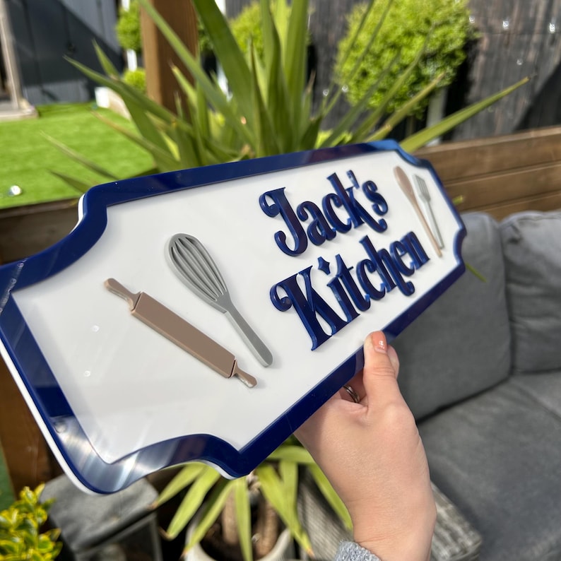 May include: White sign with a blue border and the text "Jack's Kitchen" in blue raised letters. The sign has a rolling pin and a whisk on it.