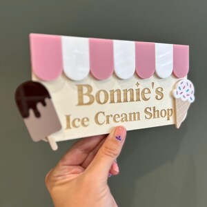 May include: A decorative sign for "Bonnie's Ice Cream Shop" featuring a pink and white striped awning design. The sign includes a chocolate ice cream bar and an ice cream cone with sprinkles. The sign is held by a hand.
