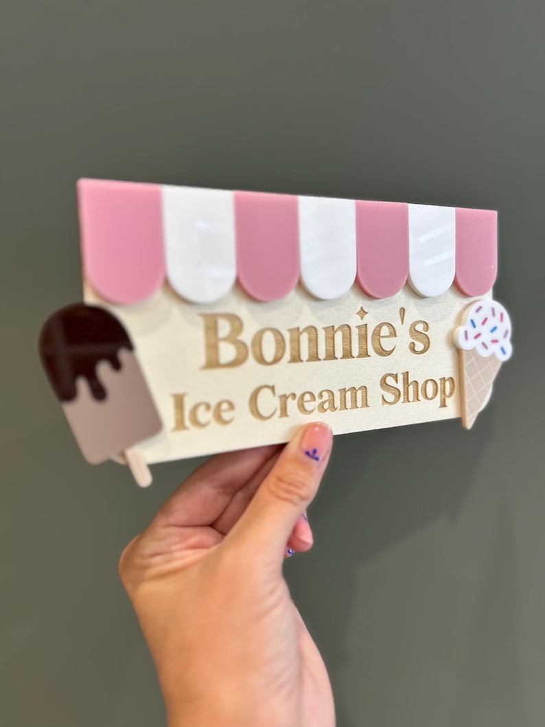 May include: A decorative sign for "Bonnie's Ice Cream Shop" featuring a pink and white striped awning design. The sign includes a chocolate ice cream bar and an ice cream cone with sprinkles. The sign is held by a hand.