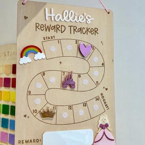 May include: A rectangular wooden reward tracker board with the text Hallie's Reward Tracker, a numbered path from 1 to 14, and a blank white reward space. It features decorative acrylic icons including a rainbow, heart, castle, crown, and princess.