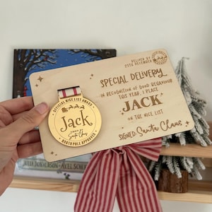 Peut inclure: Une plaque de Noël en bois avec une médaille d'or. La plaque indique "Livraison Spéciale" et "Jack sur la liste des gentils", signée par le Père Noël. La médaille dit "Prix Officiel de la Liste des Gentils" et "Jack". Un nœud rayé rouge et blanc est visible.