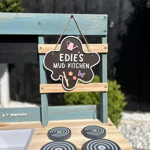 May include: A wooden play kitchen painted in blue and white. The kitchen has a sign that reads "Edie's Mud Kitchen" with a pink watering can and flowers. The kitchen has four black stovetop burners.