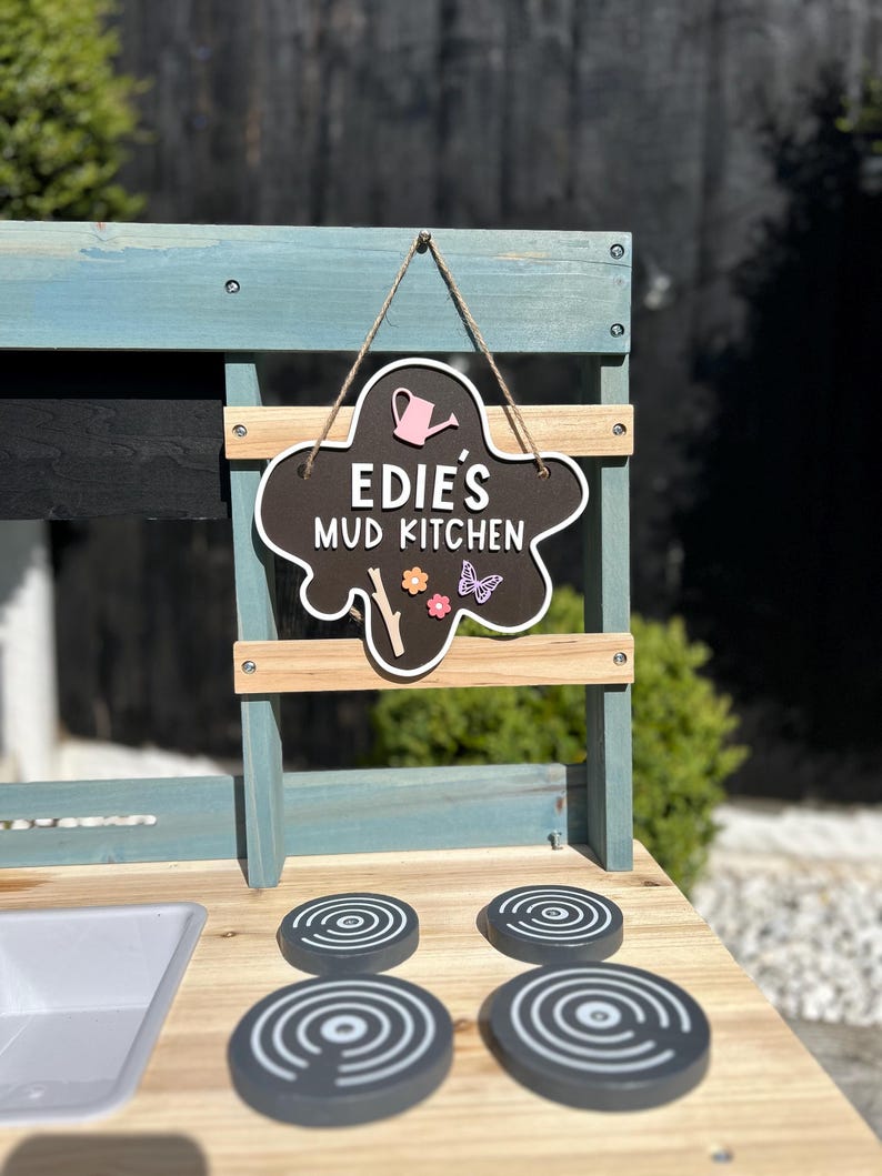 May include: A wooden play kitchen painted in blue and white. The kitchen has a sign that reads "Edie's Mud Kitchen" with a pink watering can and flowers. The kitchen has four black stovetop burners.