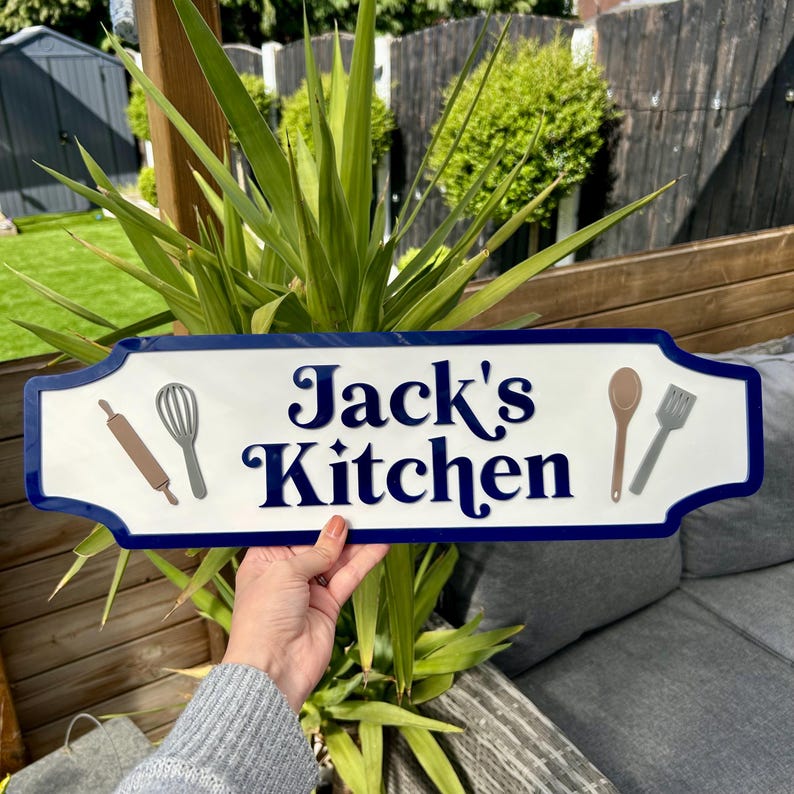 May include: White sign with blue trim and rounded edges. The sign says "Jack's Kitchen" in a decorative font. There are images of kitchen utensils on the sign, including a rolling pin, whisk, wooden spoon, and spatula.
