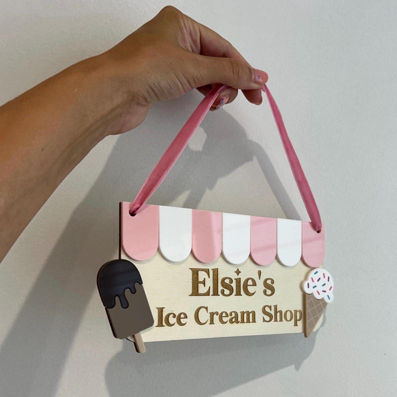 May include: Wooden sign with pink and white awning design, featuring the text "Elsie's Ice Cream Shop." Includes a chocolate ice cream bar and an ice cream cone with sprinkles. A pink ribbon is attached for hanging.