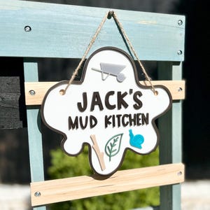 May include: White wooden sign with a brown outline in the shape of a cloud. The sign says "Jack's Mud Kitchen" with a wheelbarrow, leaf, and snail graphic.
