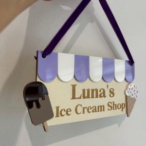 May include: Wooden sign for "Luna's Ice Cream Shop" with a purple and white awning design. Features a chocolate ice cream bar and an ice cream cone with sprinkles. The sign is suspended by a purple ribbon.