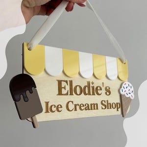 May include: Wooden sign for "Elodie's Ice Cream Shop" with a yellow and white awning design. The sign features a chocolate-covered popsicle and an ice cream cone with sprinkles. A white ribbon is attached for hanging.