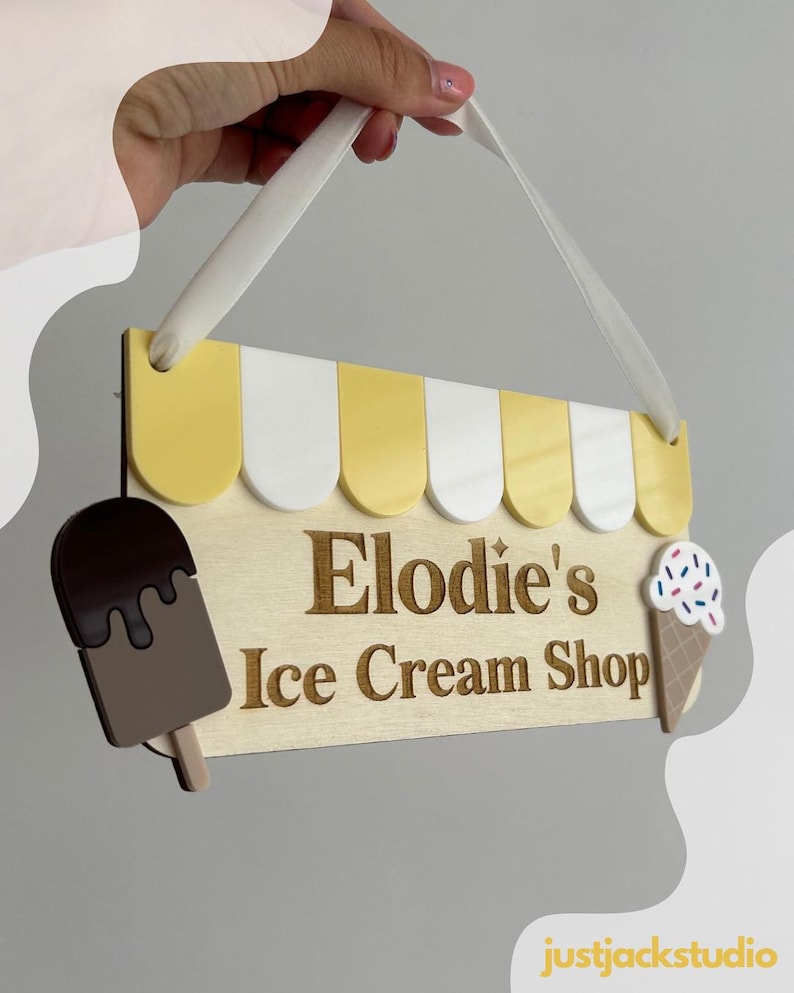May include: Wooden sign for "Elodie's Ice Cream Shop" with a yellow and white awning design. The sign features a chocolate-covered popsicle and an ice cream cone with sprinkles. A white ribbon is attached for hanging.