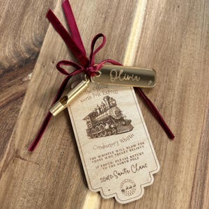 May include: A gold-toned metal whistle attached to a rectangular wooden tag and a gold acrylic name tag reading Olivia. The wooden tag features a steam train illustration and text reading The Official North Pole Express Conductor's Whistle, tied together with a dark red velvet ribbon.