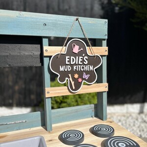 May include: A wooden play kitchen with a black chalkboard sign that says "Edie's Mud Kitchen". The kitchen has a grey sink and four black stovetop burners with white circles.