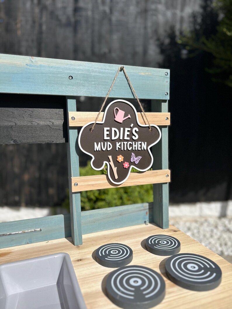 May include: A wooden play kitchen with a black chalkboard sign that says "Edie's Mud Kitchen". The kitchen has a grey sink and four black stovetop burners with white circles.