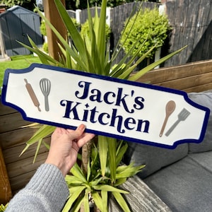 May include: White sign with blue trim and the text "Jack's Kitchen" in a blue cursive font. The sign has a rolling pin, whisk, wooden spoon, and spatula graphics.