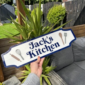 May include: White sign with blue trim and the text "Jack's Kitchen" in a retro font. The sign has a decorative border and features images of kitchen utensils, including a rolling pin, whisk, spoon, and spatula.