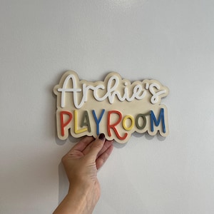 May include: A wooden sign that reads "Archie's PLAYROOM" in colorful, raised letters. The sign has a cream-colored base with the name in white script. The word "PLAYROOM" is in red, yellow, green, and blue.