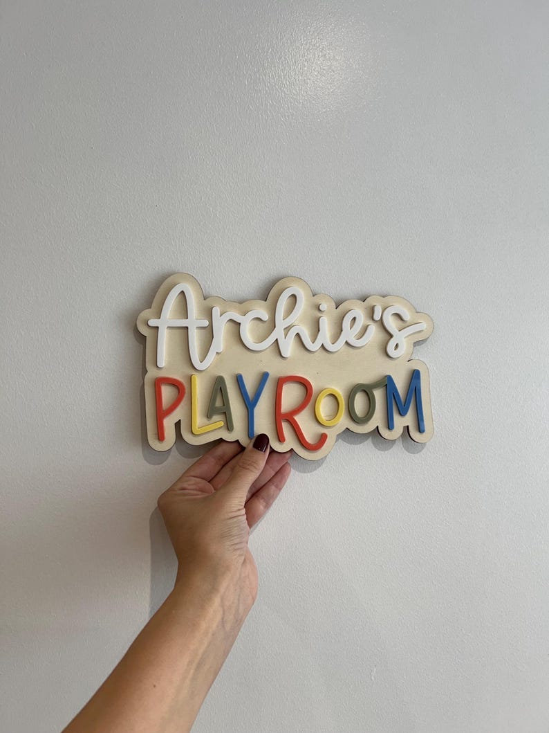 May include: A wooden sign that reads "Archie's PLAYROOM" in colorful, raised letters. The sign has a cream-colored base with the name in white script. The word "PLAYROOM" is in red, yellow, green, and blue.
