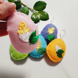 Op de afbeelding: Vijf vilten paaseieren met geborduurde kuikens en bladeren. De eieren zijn roze, groen, blauw, paars en oranje.